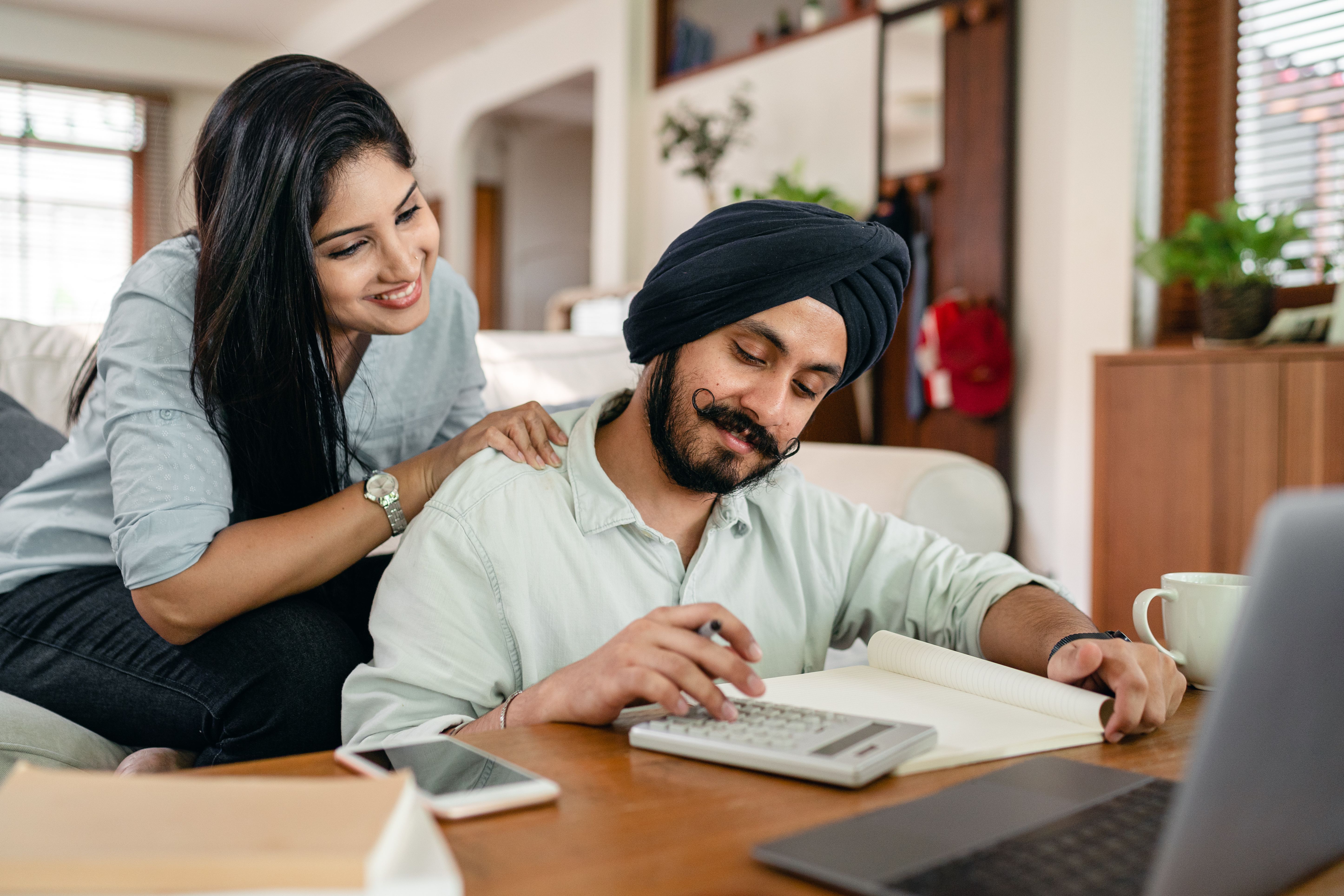Smiling ethnic woman looking at ethnic husband calculating among laptop and smartphone and books and notebook while working remotely from home